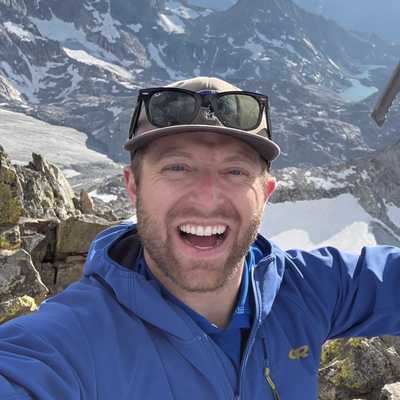 Justin grinning on a rocky summit with snowy peaks and alpine lakes behind